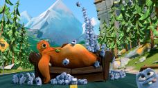 A big brown bear laying on a couch in the middle of a road with tall trees and a mountain in the background. He is surrounded by tiny, blue creatures.