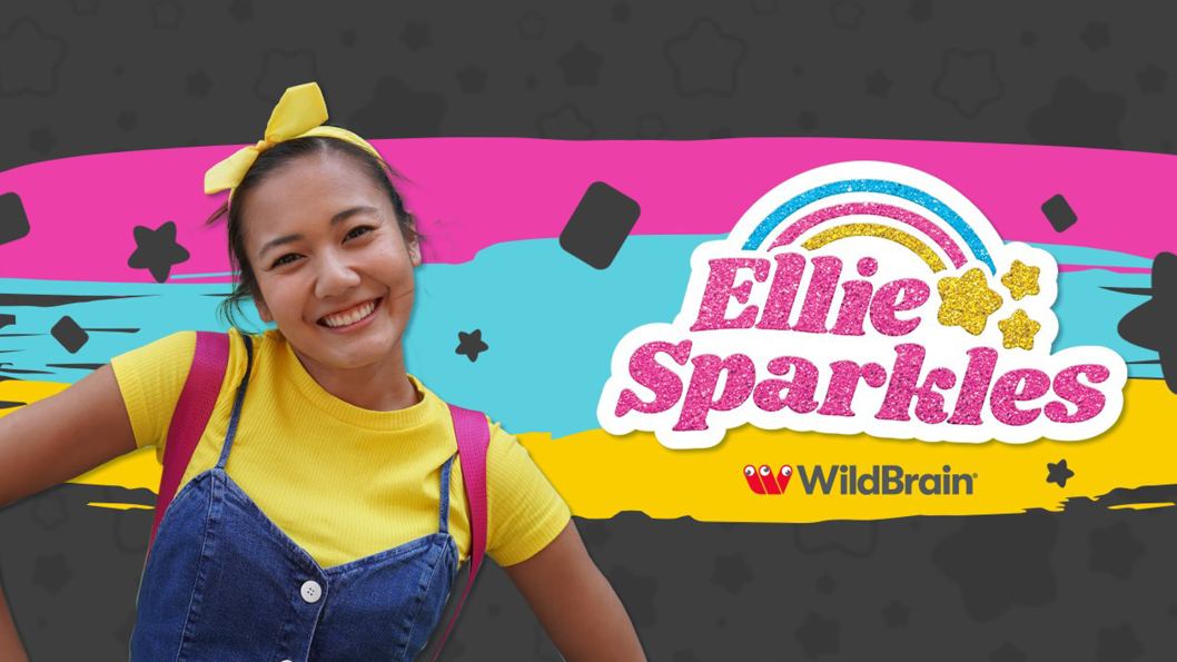 A happy girl wearing a yellow t-shirt standing in front of a colourful logo that reads, Ellie Sparkles.